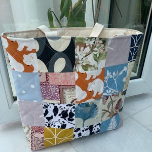 May include: A patchwork tote bag featuring diverse fabric patterns, such as polar bears, floral motifs, and geometric designs. It has cream-colored handles and a neutral interior. The bag is approximately 35 cm tall.