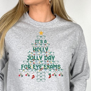 May include: Gray long-sleeve sweatshirt with a green Christmas tree design made of eyeglasses. The tree is decorated with yellow lights and red and green ornaments. The text on the sweatshirt reads "It's a Holly Jolly Day for Eye Exams."