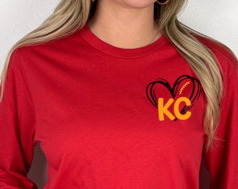 May include: A red long-sleeve shirt featuring a black heart-shaped graphic on the upper left chest. The heart design incorporates a yellow football lace pattern and the yellow letters KC centered within the heart shape.