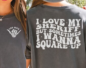 May include: A gray t-shirt with the text "I love my sheriff but sometimes I wanna square up" printed on the back in white. The front of the shirt has the text "Leo Wife" and a skeleton hand making a shaka sign.