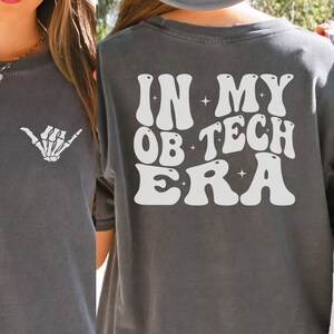 May include: A dark gray t-shirt with a white skeleton hand graphic on the front and the text "IN MY OB TECH ERA" in a retro font on the back.