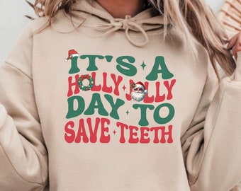May include: A beige hoodie with a green and red graphic that says "It's a Holly Jolly Day to Save Teeth". The graphic includes a Santa Claus, a wreath, and a Santa hat.