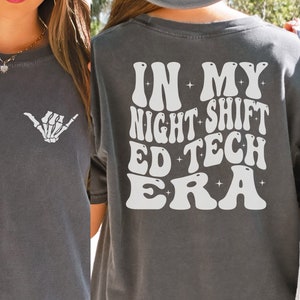 May include: A dark gray t-shirt with a white graphic on the back that says "In My Night Shift Ed Tech Era" with stars around the text. The front of the shirt has a white skeleton hand making a shaka sign.