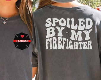 May include: Gray t-shirt with the text "SPOILED BY MY FIREFIGHTER" in white on the back. The front of the shirt has a black firefighter's badge with a red heart and the name "JACKSON" in white.