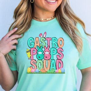 May include: A light teal t-shirt with the text "Gastro Poops Squad" in colorful bubble letters. The design includes Easter-themed graphics such as eggs, bunnies, and a chick wearing sunglasses. The shirt is a short-sleeved crew neck.