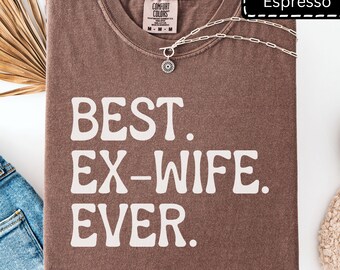 May include: An espresso brown crewneck t-shirt featuring the white text "BEST. EX-WIFE. EVER." printed on the front. The shirt has a Comfort Colors label at the inner neckline and is displayed with a silver-toned chain necklace resting on top.