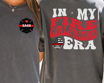 May include: Gray t-shirt with a firefighter theme. The front features a black firefighter Maltese cross with a red stripe and the name "ZACH". The back says "IN MY FIRE GIRLFRIEND ERA" in red and white, with a US flag and red line.