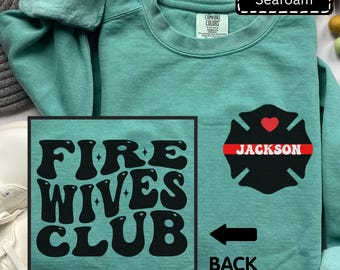 May include: A seafoam green crewneck sweatshirt featuring a black graphic on the front left chest of a Maltese cross with a red heart and the name JACKSON. The back design displays the black text FIRE WIVES CLUB in a bold, wavy font.