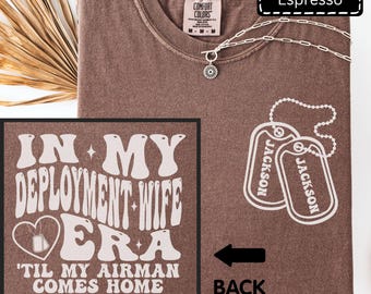 Puede incluir: Una camiseta color espresso con un gráfico que incluye el texto 'IN MY DEPLOYMENT WIFE ERA 'TIL MY AIRMAN COMES HOME' y una ilustración de dos placas de identificación con la etiqueta 'JACKSON'. La camiseta incluye un collar de cadena plateada.