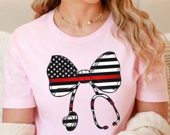 May include: A light pink t-shirt featuring a bow tie design with a red stripe and a stethoscope. The bow tie has a section with the American flag and a section with black and white stripes. The stethoscope is black and red.