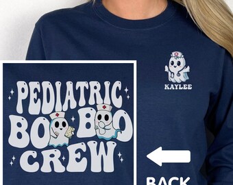 May include: Navy blue sweatshirt with the words "PEDIATRIC BOO CREW" in white, with ghost illustrations wearing nurse hats. A smaller ghost design with the name "KAYLEE" is on the upper right. The back of the shirt is indicated by an arrow.