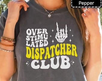 May include: Dark gray t-shirt with the text "OVER STIMULATED DISPATCHER CLUB" in white and yellow. A skeleton hand makes a rock and roll gesture. The word "Pepper" is in a white box in the upper right corner.