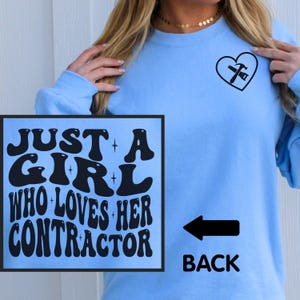 May include: Light blue sweatshirt with the text "JUST A GIRL WHO LOVES HER CONTRACTOR" on the front. A black arrow points to the back, where a heart with a hammer and saw graphic is located. The sweatshirt is a crew neck style.