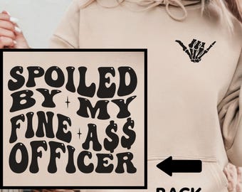 May include: Beige hoodie with the text "SPOILED BY MY FINE A$$ OFFICER" in black. A black arrow points to the right, with the word "BACK" below it. A black skeleton hand gesture is on the upper right.