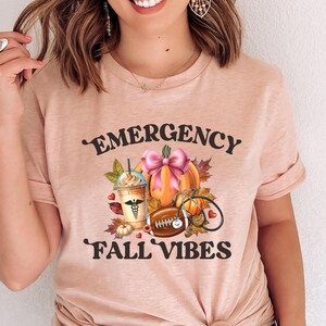 May include: A light pink t-shirt with the words "EMERGENCY FALL VIBES" in black script. The graphic includes a pumpkin with a pink bow, a coffee drink, a football, and a stethoscope.
