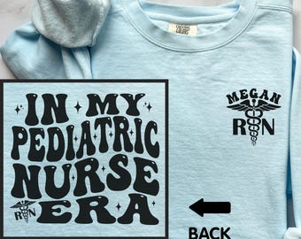 May include: Light blue sweatshirt with the text "IN MY PEDIATRIC NURSE ERA" in black, along with a medical symbol and the text "Megan RN". The back of the sweatshirt is indicated with an arrow and the word "BACK".