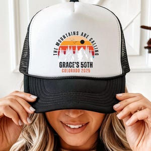 May include: A black and white trucker hat with the text "THE MOUNTAINS ARE CALLING" and "GRACE'S 50TH COLORADO 2025" in a circular design. The design features a sunset over a mountain range. The hat is being held up by a person.