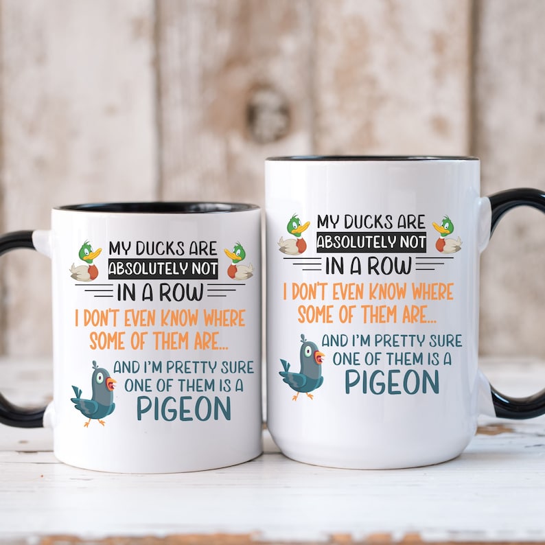 May include: Two white coffee mugs with black rims and handles. Both mugs feature a humorous graphic with ducks and a pigeon. The text on the mugs reads "My ducks are absolutely not in a row. I don't even know where some of them are... And I'm pretty sure one of them is a pigeon."