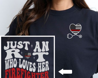 May include: A navy blue t-shirt with the text "Just an R.N. who loves her firefighter" in white and red lettering. A red line runs through a white heart with an American flag design. The back of the shirt has a stethoscope and a heart with an American flag design.