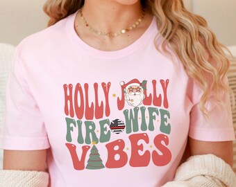 May include: A light pink t-shirt with the words "HOLLY JOLLY FIRE WIFE VIBES" in red and green retro font. The design includes a Santa Claus illustration, a Christmas tree, and an American flag.