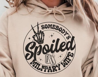 May include: Beige hoodie with a circular graphic. The graphic features the text "SOMEBODY'S Spoiled MILITARY WIFE" in a stylized font, along with a skeleton hand making a peace sign and two dog tags.