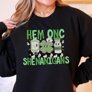 May include: Black sweatshirt with a green and white graphic. The graphic includes the words "HEM ONC SHENANIGANS" above cartoon images of a medical IV bag, a four-leaf clover, and a medicine bottle.