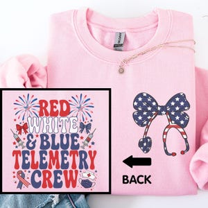 May include: A light pink sweatshirt with a graphic on the front that says "RED WHITE & BLUE TELEMETRY CREW". The back of the sweatshirt has a stethoscope design with a bow made of stars and stripes. A necklace with a small pendant is also visible.