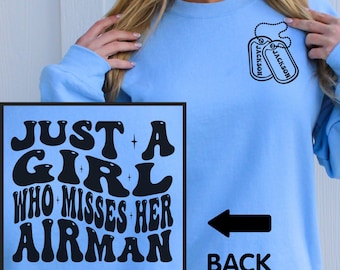 May include: A light blue crewneck sweatshirt featuring a black graphic on the chest of two dog tags labelled JACKSON. The back of the sweatshirt displays the black text JUST A GIRL WHO MISSES HER AIRMAN in a bold, wavy, retro-inspired font.