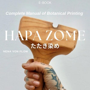 May include: A wooden mallet with a handle, used for hammering fabric in the Japanese textile dyeing technique called "Tatakae Zome". The text "Tatakae Zome" is written in Japanese characters on the mallet. The mallet is held by a hand wearing a white fabric with a blue floral pattern.
