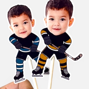 May include: Two die-cut paper toppers featuring a young child's face on a cartoon hockey player body. One figure wears a black and teal uniform with teal socks, while the other wears a black and gold uniform with gold striped socks, both holding sticks.