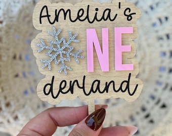 May include: A wooden cake topper with a scalloped edge featuring the black text Amelia's and derland, a silver glittery snowflake, and the pink letters NE. The topper is attached to a thin wooden stick, creating a decorative sign for a celebration.
