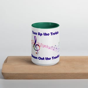 Op de afbeelding: Witte keramische mok met een groene rand. De mok heeft een kleurrijke vioolsleutel en muzieknoten met de tekst "Turn Up the Treble Drown Out the Trouble" in paars.