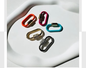 May include: An assortment of colorful oval-shaped carabiner clasps in orange, pink, teal, gold, and silver. The clasps are displayed on a white, curved tray. Some clasps have decorative embellishments.