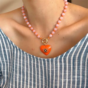 May include: A pink and orange beaded necklace with a gold heart pendant featuring an evil eye design.