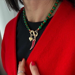 May include: A beaded necklace featuring round green beads interspersed with small orange beads. The necklace includes a gold-toned metal clasp at the center, from which hang a small gold-toned heart charm and a red chili pepper-shaped pendant.