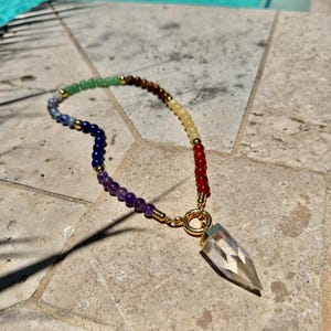 Peut inclure: Un collier de perles colorées avec un pendentif en cristal transparent. Les perles sont disposées en arc-en-ciel, avec des accents dorés. Le pendentif est un cristal clair et pointu. Le collier est présenté sur une surface en pierre.