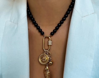 May include: A necklace featuring a strand of smooth black beads connected to a gold-toned oval carabiner clasp. The clasp is embellished with small clear stones and holds three gold-toned charms: a circular lion head, a small sphere, and a fish.