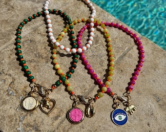 May include: Four beaded bracelets in green, white, yellow, and pink. Each bracelet has a gold clasp and a charm. Charms include a coin, heart, pink medallion, seashell, evil eye, and elephant.