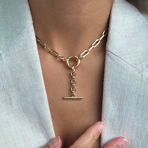 Op de afbeelding: Goudkleurige ketting met een toggle sluiting. De ketting heeft een schakelketting ontwerp met een cirkelvormige hanger en een verticale staafsluiting. De sieraden worden gedragen over een lichtgekleurde blazer.