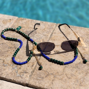 Puede incluir: Gafas de sol doradas con lentes ovaladas y oscuras, sobre una superficie de piedra. Las gafas están adornadas con una cadena de cuentas verdes y azules, y detalles de cadena dorada. En el fondo, una piscina.