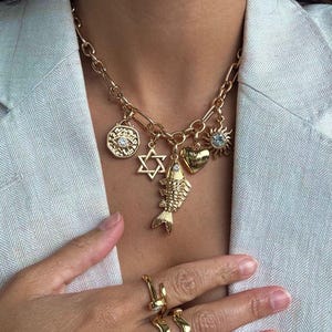 May include: Gold-toned charm necklace featuring a variety of pendants, including a fish, Star of David, heart, sun, and an eye. The necklace is paired with gold rings. The jewelry is displayed against a light-colored blazer.