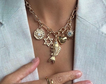 May include: Gold-toned charm necklace featuring a variety of pendants, including a fish, Star of David, heart, sun, and an eye. The necklace is paired with gold rings. The jewelry is displayed against a light-colored blazer.