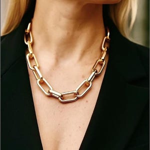 May include: A chunky gold chain necklace with large, rectangular links. The necklace is worn over a black blazer. The jewellery is a striking statement piece, with each link clearly visible.
