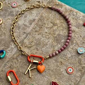 Puede incluir: Un collar con una cadena dorada en un lado y una hilera de cuentas de color rosa empolvado en el otro. Un mosquetón naranja une ambos lados, sosteniendo un dije con la letra K dorada y un pequeño dije en forma de corazón naranja.