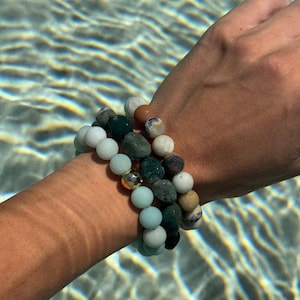 May include: Three beaded bracelets in shades of green, white, and brown are stacked on a wrist. The bracelets feature a variety of stone beads, including round and irregular shapes. A gold-colored bead accents one bracelet. The background is a body of water.