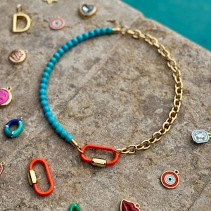 May include: A necklace featuring a half-strand of turquoise beads and a half-strand of gold-tone chain, connected by a bright orange carabiner clasp. Several small charms, including a letter D, a letter B, a red lip, and an eye, are scattered nearby.