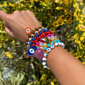 May include: A collection of beaded bracelets in various colors, including red, blue, orange, and white. Some bracelets feature eye-shaped charms and heart-shaped beads. The bracelets are stacked on a wrist.