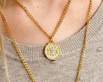 May include: Gold-coloured necklace with an eye pendant, featuring a small, clear stone. The necklace has a layered design with a thicker chain and a thinner chain. The background is a grey jumper.