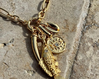 May include: Gold-toned necklace with a variety of charms, including a fish, a horn, a coin with an eye design, and a shell. The chain is a chunky link style, and the charms are detailed with small embellishments.