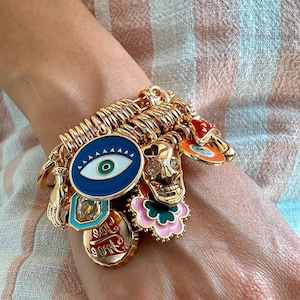 Può includere: Un bracciale con ciondoli color oro dotato di vari pendenti, tra cui un occhio blu, un teschio metallico, un fiore rosa e verde, un fungo rosso e bianco e un ciondolo circolare con la scritta "Too Free" in corsivo rosso.